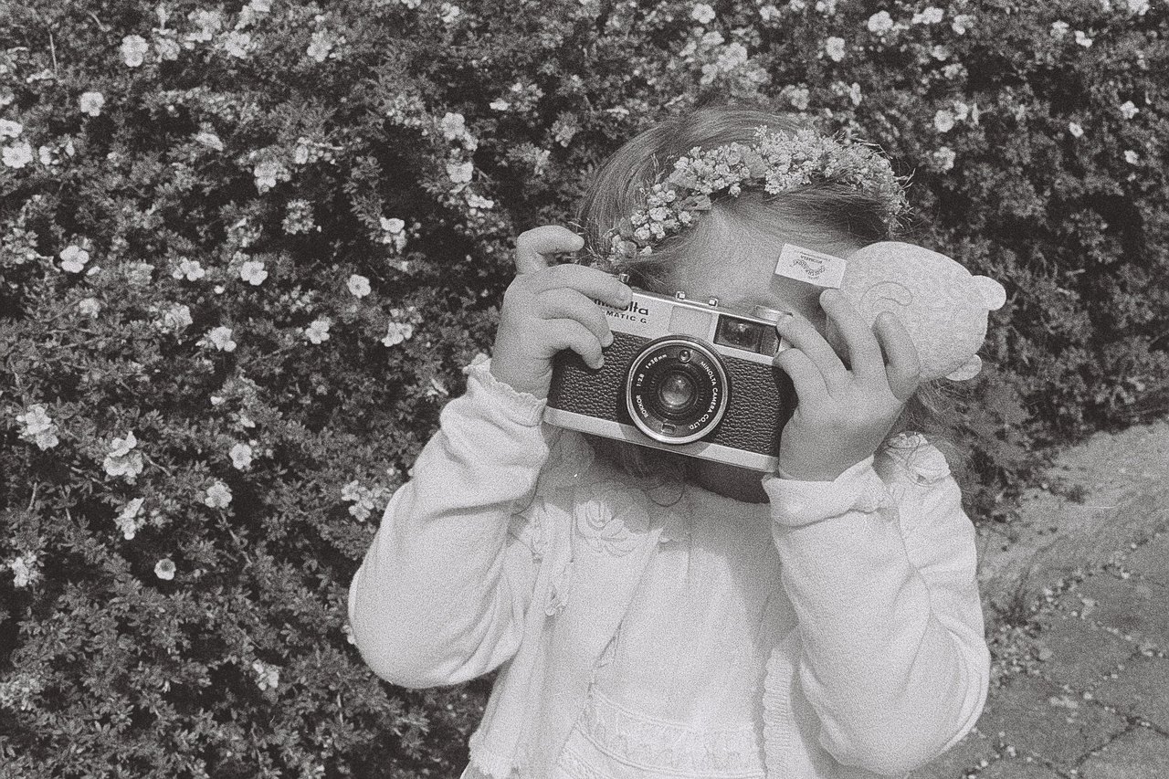 A small child in a white cardigan and a flower crown holds a rangefinder camera up to her face, in front of a flowering hedge, shot on black and white 35mm film