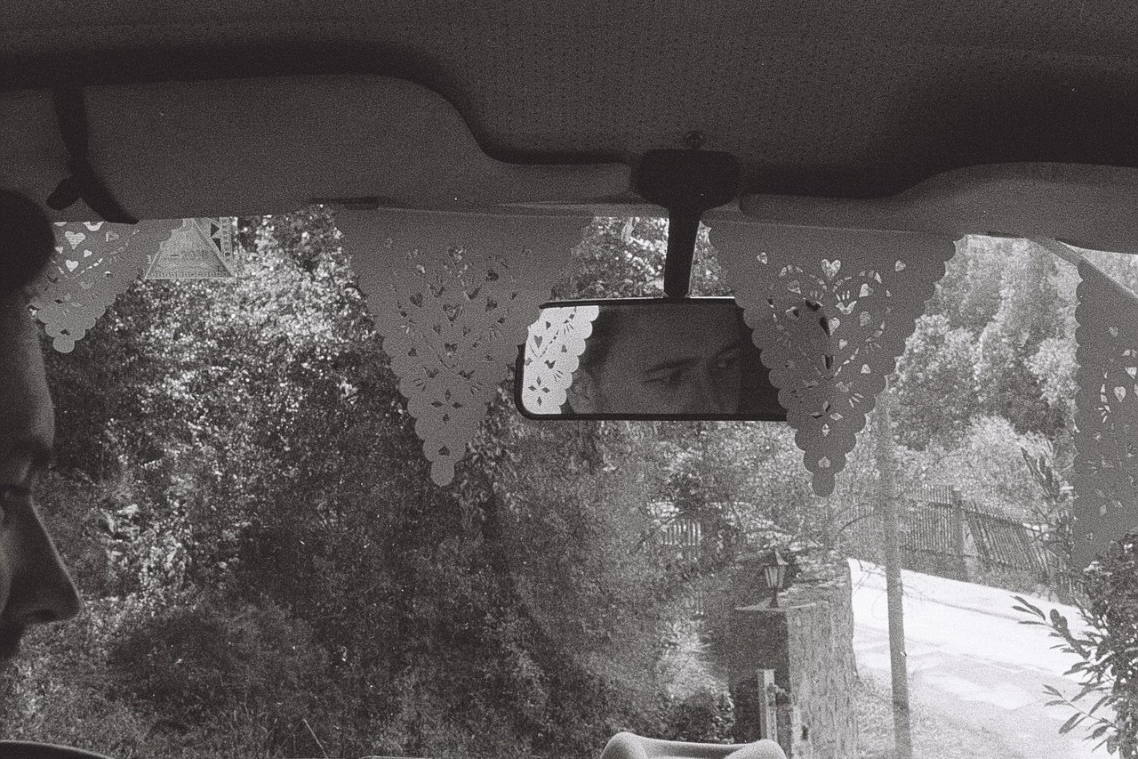 View through a car windscreen hung with paper bunting, the driver's eyes in the rear-view mirror, a leafy road ahead, shot on black and white 35mm film